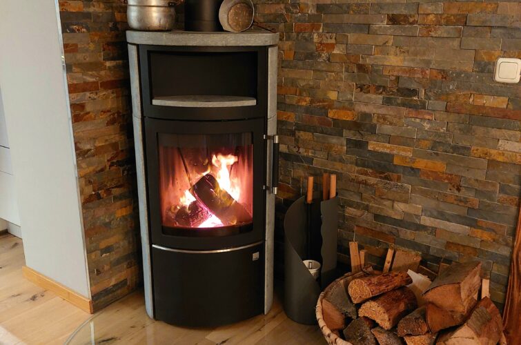 Holzofen Hark 44 5.1 in schwarz / Speckstein im Wohnzimmer. Darauf steht eine alter Topf und daneben ein Holzkorb.
