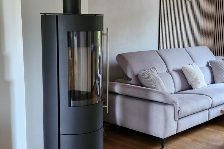 Holzofen Olsberg Tacora in anthrazit in einem Wohnzimmer mit Holzboden. Daneben steht ein helles Sofa.